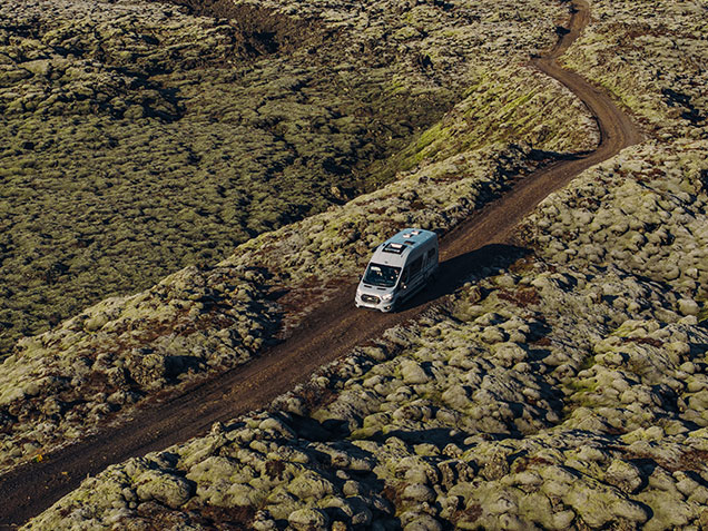 Camping auf Island: Campervan fährt auf einer nicht asphaltierten Straße inmitten mattgrüner Mooslandschaften entlang.