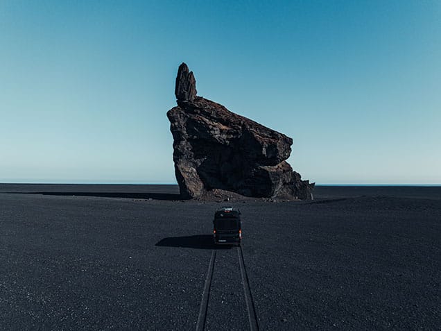 Camping auf Island: Campervan fährt auf schwarzem Sand auf einen dunklen Felsen zu.