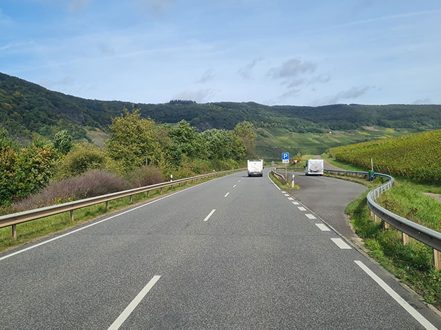 Blick aus einem Fahrzeug auf eine Straße an der Mosel mit vorausfahrenden Wohnmobilen, umgeben von grüner Natur.