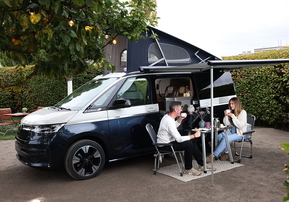 California Campervan mit zwei Personen auf Campingstühlen im California Garden in der Autostadt Wolfsburg.